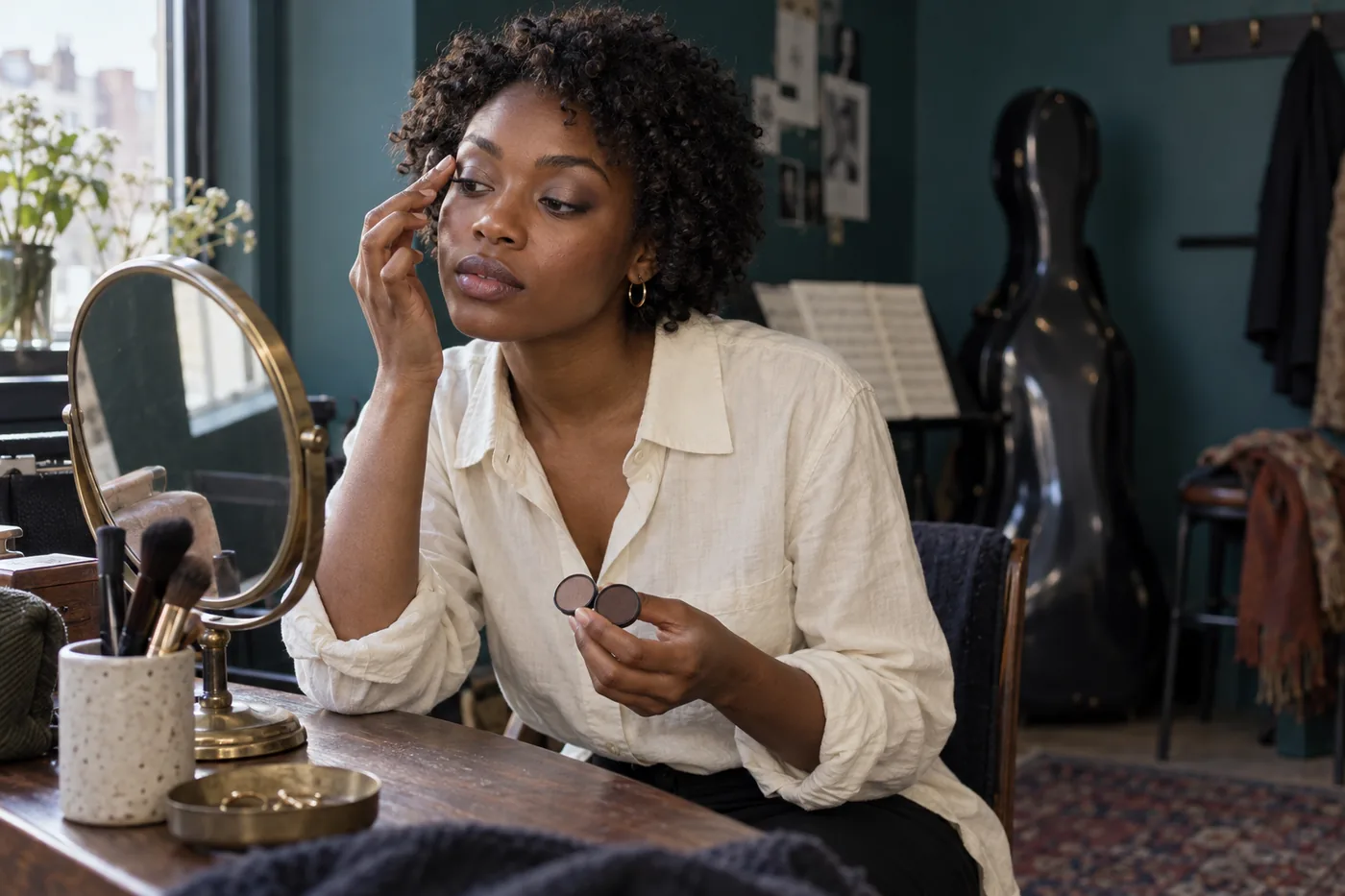 Young Black adult with natural curls checks taupe eye makeup beside a mirror in a teal rehearsal dressing room