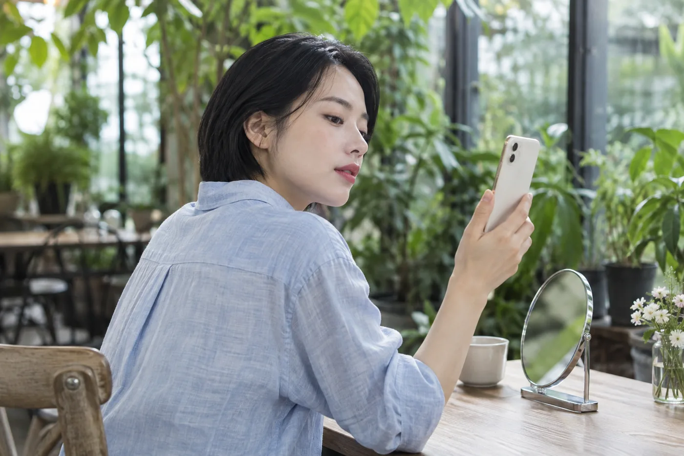 Young East Asian adult in a pale blue shirt looks at a phone beside a small mirror in a leafy greenhouse cafe
